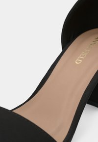 Mustassa korkokenkä, jossa on samettipinta, avokärkinen muotoilu ja sileä beige pohja, jossa on kultainen brändäysdetalji.