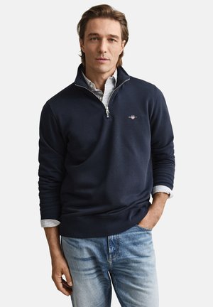 Marineblauwe sweatvest met ritssluiting, geribbelde manchetten en tailleband, gedragen over een lichtblauw gestreept overhemd met knoopsluiting, gecombineerd met lichte denim jeans.