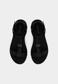 Μαύρα σανδάλια με επίπεδες σόλες, με ρυθμιζόμενους ιμάντες και χρυσή λεπτομέρεια στο κέντρο. Λείο συνθετικό υλικό με μινιμαλιστικό σχεδιασμό.