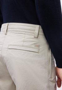 Close-up of beige pants pocket with BOSS label worn with a black long-sleeve top, showing stitching and belt loops, hand resting by side.