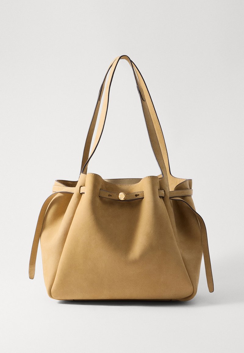 Tan suède tote bag met platte schouderbanden, een geraffineerde bovenkant met trekkoordsluiting en een gouden knopdetail. Gladde textuur en minimalistisch ontwerp.