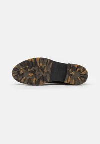 Suela de zapato de goma negra con patrón de agarre texturizado, con acentos en camuflaje marrón y amarillo. Diseñada para durabilidad y tracción.