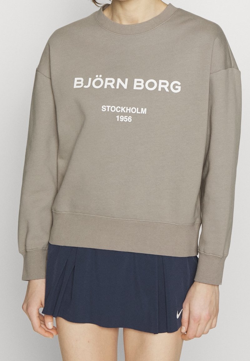 Personne portant un sweat-shirt Björn Borg taupe avec le texte "STOCKHOLM 1956" et une jupe plissée bleu marine avec un petit logo blanc Nike.