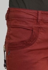Red cotton blend pants with a smooth texture, featuring a curved pocket design and decorative stitching along the pocket edges.