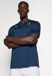 Marineblaue kurzarm Sportshirt aus strukturiertem Stoff, mit Rundhalsausschnitt und Schnürdetail. Logopatch auf der Brust, gerader Saum.