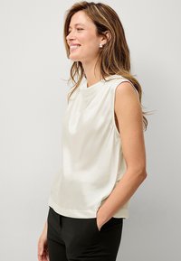 Ärmelloses Seidenoberteil in Off-White mit einem drapierten Ausschnitt und seitlichen Details, kombiniert mit schwarzen Hosen. Glatte Textur und subtiler Glanz.