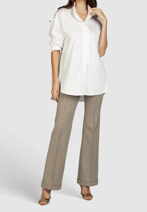 Femme portant un chemisier blanc à manches longues, un pantalon beige évasé à motifs et des talons ouverts dorés, debout devant un fond uni.