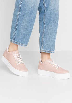 Osoba ubrana w jasnoniebieskie dżinsy i różowe sneakersy na platformie ze sznurowadłami stoi na białej podłodze.