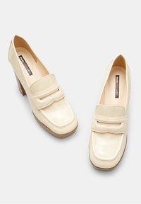 Mocassins en cuir verni crème avec un talon carré, dotés d'un bout arrondi et d'une bride décorative sur le coup de pied. Semelle en caoutchouc texturée.