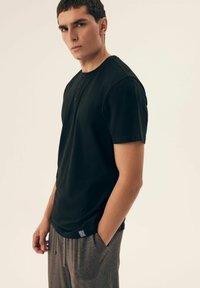 Black short-sleeved henley shirt with three button placket. Fabric has a smooth texture. Worn with gray drawstring pants featuring logo detail.