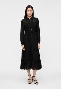 Μαύρο φόρεμα με μακριά μανίκια, εμπρός με κουμπιά, με σμιλευμένο κορσέ και τελείωμα με βολάν σε στρώσεις. Κατασκευασμένο από ελαφρύ, διαφανές ύφασμα.