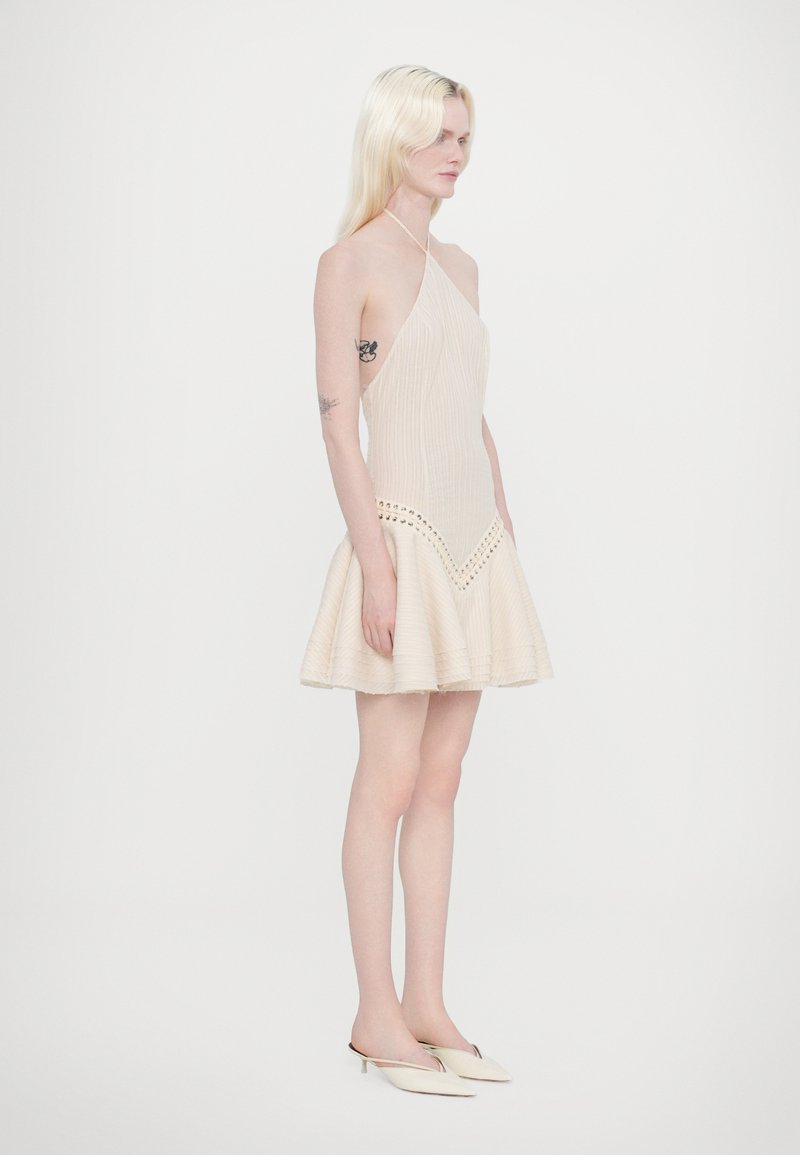 Beige pleated mini dress with an asymmetrical halter neckline, featuring a scalloped hem and decorative cut-outs near the waist.
