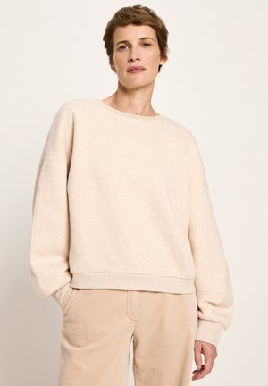 Eine Frau trägt einen lockeren beigen Sweatshirt und beigen Cordhosen und steht mit einer Hand in der Tasche vor einem schlichten hellen Hintergrund.