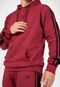 Burgundowa bluza z kapturem z czarnymi paskami akcentującymi, z kapturem na sznurku, kieszenią z przodu oraz haftowanym logo na klatce piersiowej. Wykonana z miękkiej, teksturowanej tkaniny.