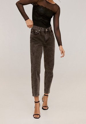 Mujer con blusa negra transparente de manga larga, jeans negros de tiro alto lavados y sandalias negras de tacón alto con correa en el tobillo, de pie.