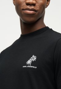 Κοντινό πλάνο ενός ατόμου που φοράει μαύρο πουκάμισο με λευκά γραφικά φοίνικα και το κείμενο "KARL LAGERFELD" στο στήθος.