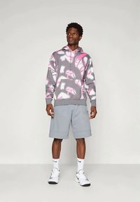 Γκρι φούτερ με κουκούλα, διακοσμημένο με ροζ και λευκό αφηρημένο μοτίβο, συνδυασμένο με ελαφριά γκρι σορτς. Τα παπούτσια είναι λευκά με μαύρες λεπτομέρειες και οι κάλτσες είναι λευκές.