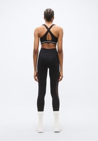 Μαύρο αθλητικό σουτιέν με λουριά που διασταυρώνονται στην πλάτη, μαύρο κολάν με ψηλή μέση και διακοσμητικές διαφάνειες στα πλαϊνά, συνδυασμένο με λευκές αθλητικές κάλτσες και παπούτσια.