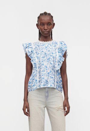 Kobieta z warkoczami, ubrana w białą bluzkę z niebieskim kwiatowym wzorem i falbankowanymi rękawami, w zestawieniu z jasnymi, podartymi jeansami.