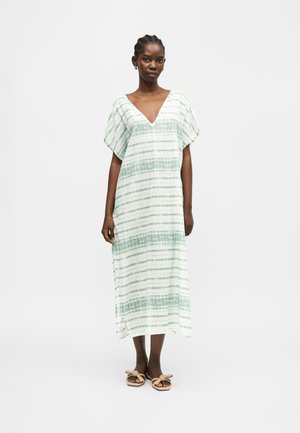 Naine kannab valget ja helerohelise triibulist lahtist maksikleiti lühikeste varrukatega ning sandaale paeladega, seisab valgel taustal.