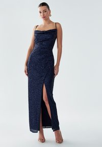 Navy sequined evening dress with a cowl neckline, thin straps, and a thigh-high slit. Smooth texture, floor-length design, fitted silhouette.