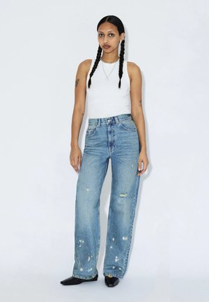 Kvinde med langt fletning, iført en hvid ærmeløs top, højttaljede, falmede blå jeans, sorte spidse sko og sølvsmykker, stående.