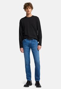 Camicia nera a maniche lunghe, jeans di denim blu con taglio dritto, cintura nera e scarpe nere stringate indossate da un modello su uno sfondo neutro.