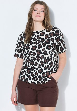 Kobieta w bluzce z krótkim rękawem w panterkę i ciemnobrązowych krótkich spodenkach, stojąca z jedną ręką w kieszeni na tle gładkiej białej ściany.