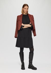 Czerwono-czarny żakiet w wzór houndstooth na czarnej sukience z warstwowym dołem, w połączeniu z czarnymi botkami do kolan i czarnymi rajstopami.
