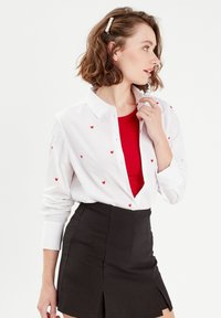 Chemise blanche à boutons avec broderie de cœur rouge, portée ouverte sur un top côtelé rouge, associée à une mini-jupe noire. Lignes épurées et texture lisse.