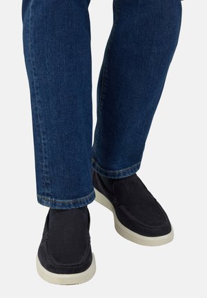 Zapatillas de ante negro sin cordones con suela de goma blanca, combinadas con unos jeans de mezclilla azul, mostrando un ajuste relajado y un corte recto.