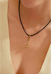 Collier en cuir noir avec un pendentif en forme de lettre « J » tordu en or, doté d'une texture lisse et d'un design minimaliste.