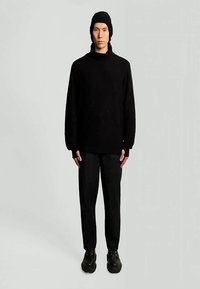 Melnas adītas džemperis ar augstu apkakli un dimanta rakstu; saskaņots ar melniem šauriem biksēm un melniem masīviem apaviem. Garas piedurknes.