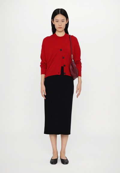 Cardigan rouge avec des boutons noirs sur une robe midi ajustée noire. Le mannequin porte des ballerines noires et porte un petit sac bordeaux. Design simple, texture lisse.