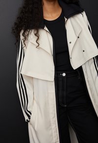 Krämfärgad jacka med lager-på-lager svart krage, med svarta ränder på ärmarna och tryckknappar. Bärs över en svart topp och jeans.