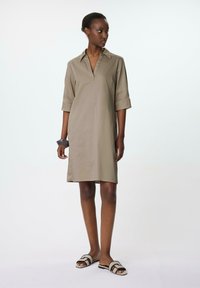 Knie-langes, hellbraunes Kleid mit Kragen und kurzen Ärmeln. Aus glattem Stoff gefertigt, mit einer Vorderblende. Getragen mit schwarzen und beigen Sandalen.