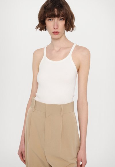Weißes Rippentop mit dünnen Trägern, kombiniert mit hochgeschnittenen beigen Hosen mit Falten und seitlichen Taschen. Glatte Textur.