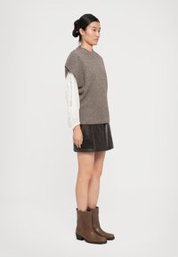 Frau mit schwarzem Haar im Dutt, die einen taupefarbenen, gestrickten ärmellosen Pullover über einer weißen Bluse, einen dunkelbraunen Lederrock und braune Stiefel bis zur Mitte der Waden trägt und seitlich steht.