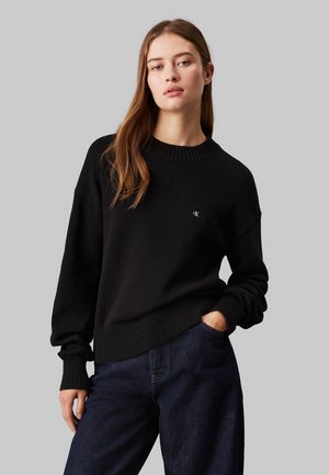 Maglione nero a maglia con collo e polsini a coste, con un piccolo logo sul petto. Indossato con jeans in denim scuro.