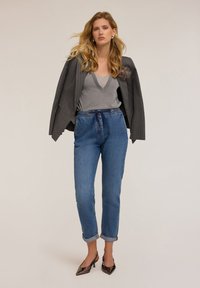 Sacou gri cu dungi subțiri peste un top gri strălucitor, asortat cu blugi deschiși la culoare cu manșete și pantofi cu toc înalt, subțiri, maro. Păr lung și ondulat.