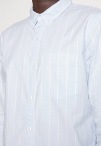 Jasnoniebieska koszula na guziki z białymi pionowymi paskami, z pojedynczą kieszenią na piersi oraz miękkim, teksturowanym materiałem.