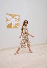 Femme en robe fleurie beige marchant à l'intérieur devant une œuvre d'art murale abstraite dorée et blanche sur un sol en bois clair.