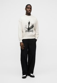 Mann iført en hvit genser med en svane og "Copenhagen"-trykk, svarte vide bukser og svart-hvite loafers, stående mot en ensfarget bakgrunn.