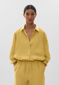 Chemise boutonnée en lin jaune avec une poche, coupe décontractée et manches retroussées, associée à un pantalon à taille élastique assorti.