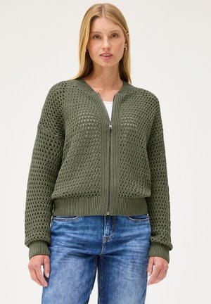 Frau trägt einen olivgrünen, offen gestrickten Zip-Pullover über einem weißen Top mit blauen Jeans vor einem einfarbigen hellen Hintergrund.