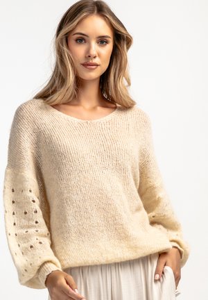 Beżowy sweter z dzianiny o luźnym kroju, z szerokim dekoltem oraz teksturowanymi rękawami z małymi wzorami wyciętymi. Miękki, puszysty materiał.