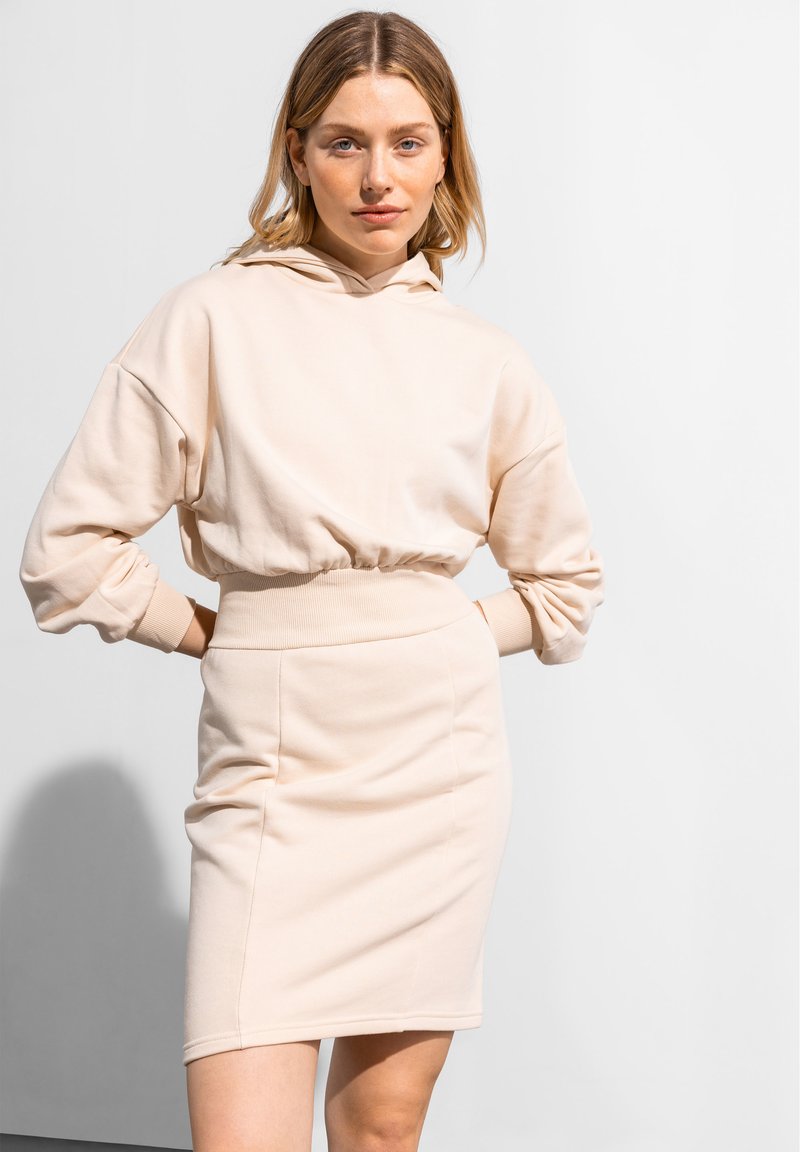 Beige Baumwoll-Hoodie mit Kordelzug und gerippten Bündchen, kombiniert mit einem taillierten beige Minirock, der eine glatte Textur und ein schlichtes Design zeigt.