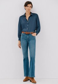 Camicia button-up in satin blu scuro con colletto, abbinata a jeans chiari a zampa d'elefante e stivaletti con tacco in camoscio marrone. Cintura marrone che mette in risalto il punto vita.