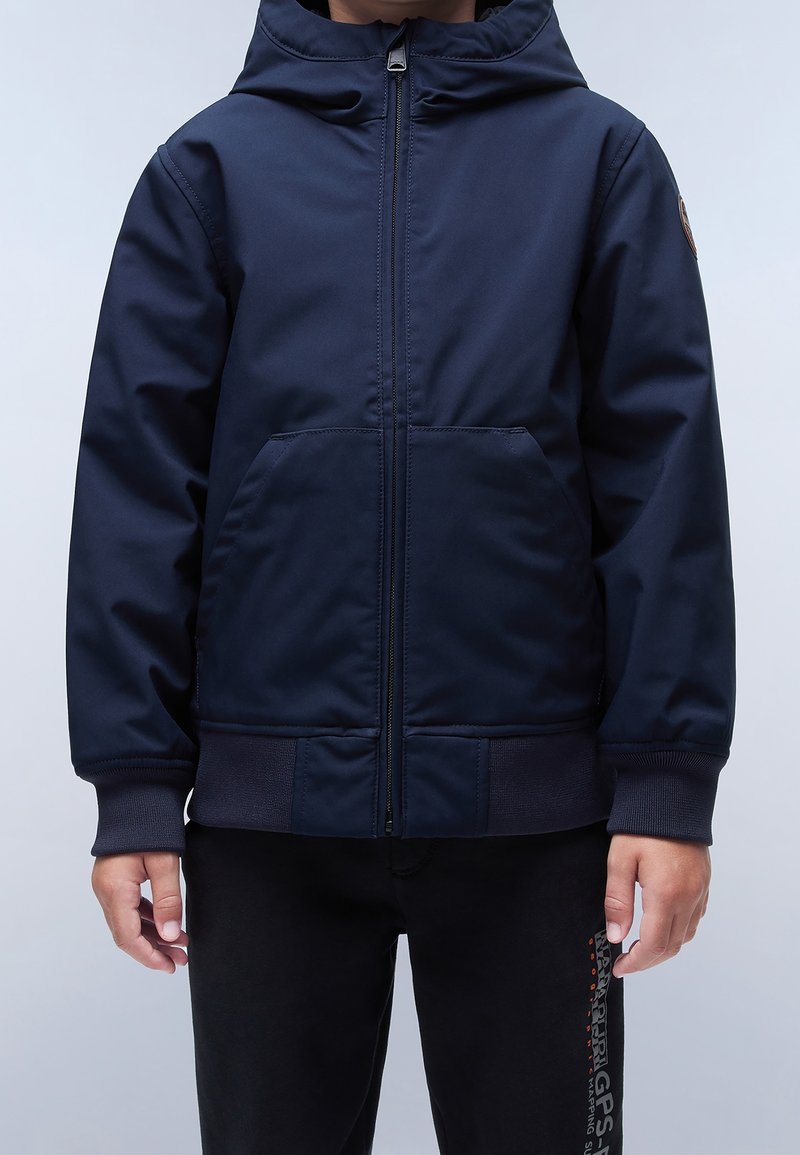 Enfant portant une veste à capuche bleu marine zippée avec poches avant et un pantalon noir avec un logo sur la jambe droite, se tenant devant un fond uni.