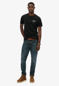 Czarny bawełniany t-shirt z białym logo, zestawiony z niebieskimi dżinsami i brązowymi butami na sznurówki, z podwiniętymi rękawami.